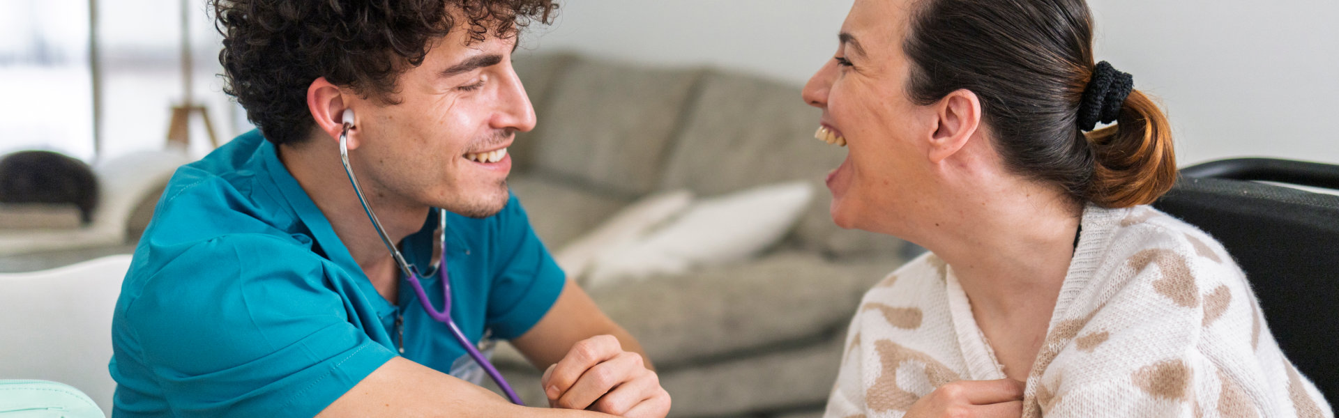 Healthcare professional with a stethoscope smiles while attending to a laughing patient in a cozy home setting.